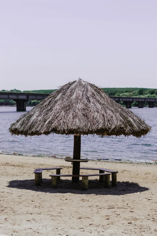 a wooden table set with a straw umbrella and round benches on a beach near the ocean