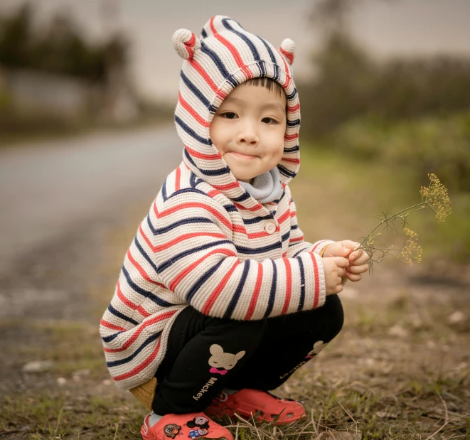 a little boy wearing a striped hooded jacket and kneels on the ground with flowers in his hand