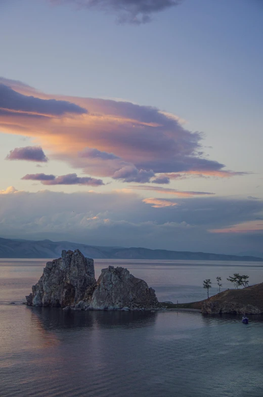 a lone rock in the water at twilight with clouds above it