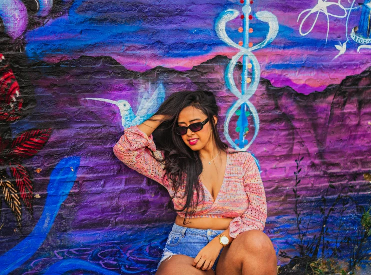 young woman wearing blue denim shorts poses next to colorful wall