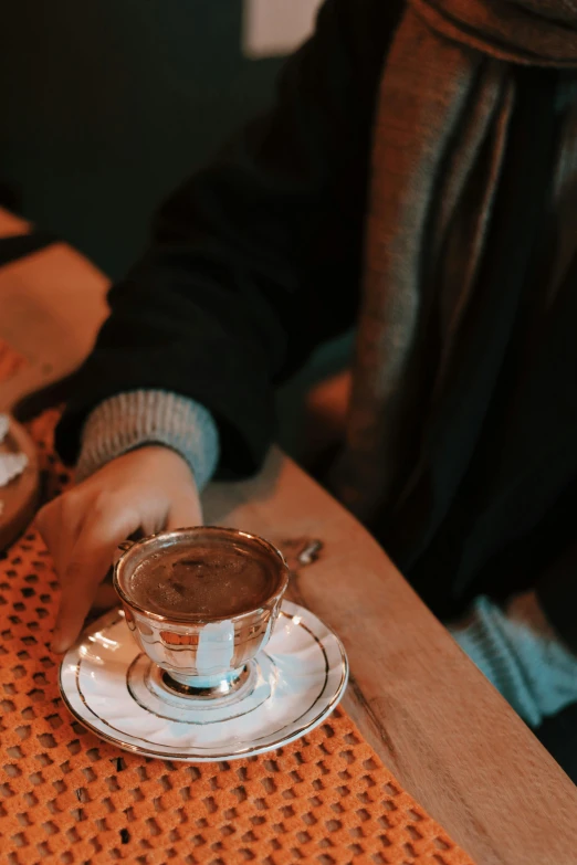 a cup of  chocolate sits on a saucer with someone holding a small plate in the background