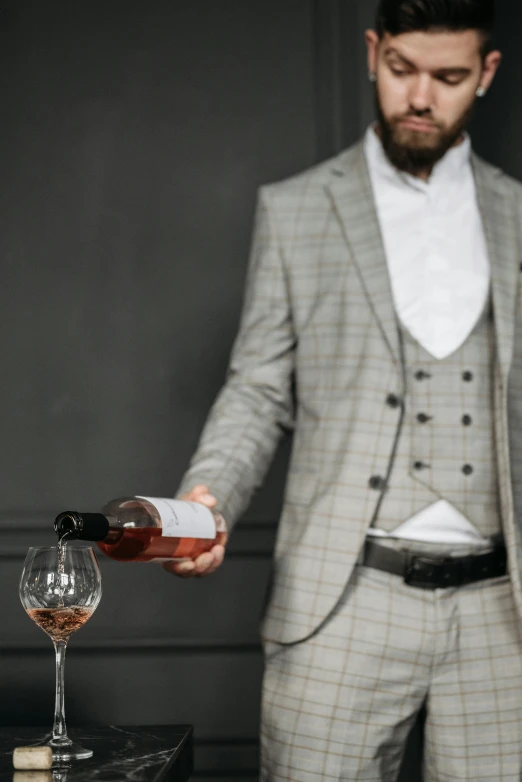 a man wearing a gray suit pouring some wine in to a glass