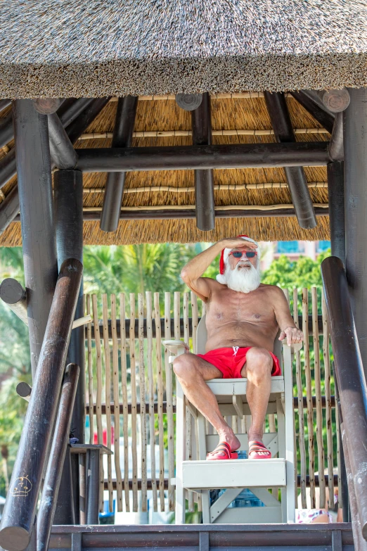 an older man in red swim trunks sits on a chair with straw covered roof