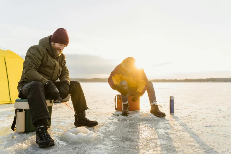 a man sitting next to a person in winter clothes and boots on a ice covered lake