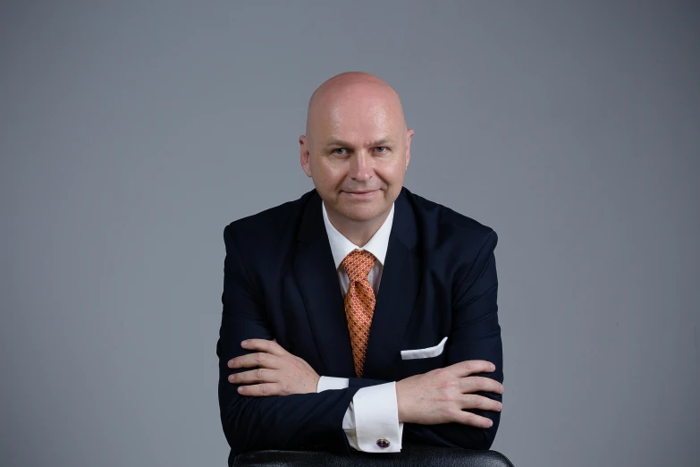 a balding man wearing a suite with a tie and sitting in front of a gray background