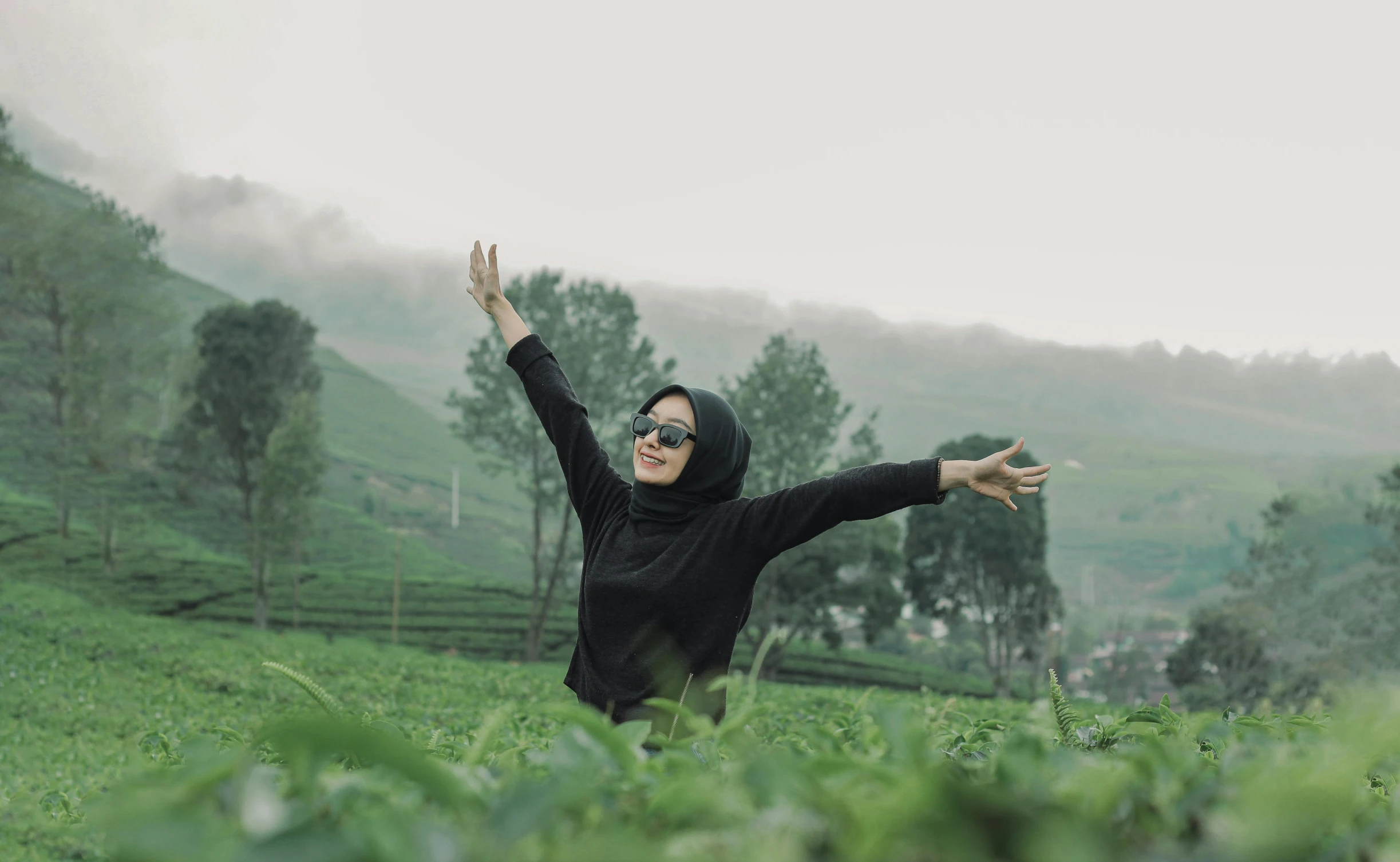 a woman in black top with her arms raised standing on grassy hill