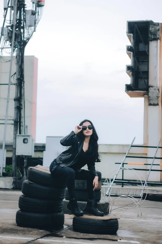 a woman in a black leather jacket and black boots sits on top of stacking tires