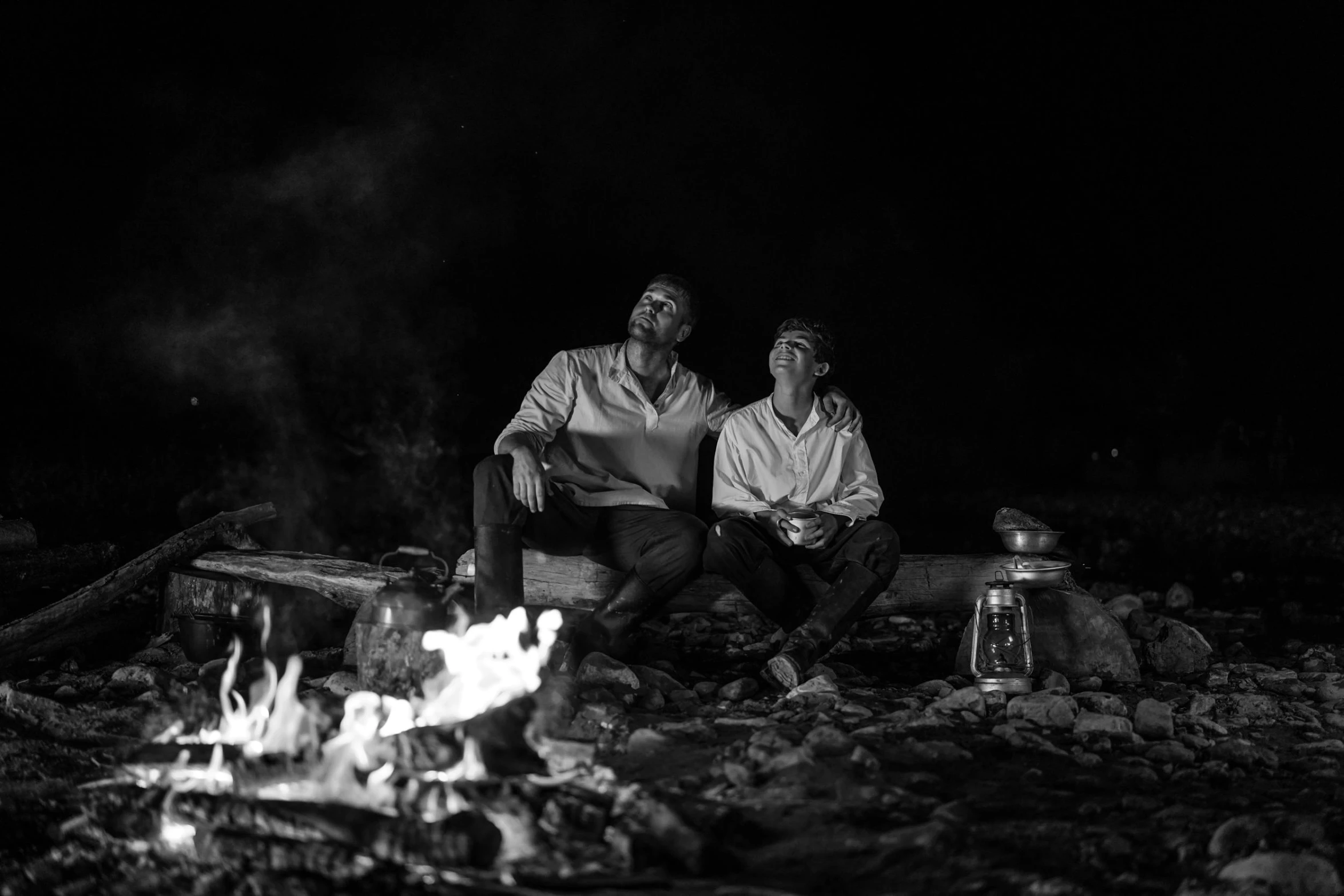 two men sit near a campfire and cook some food