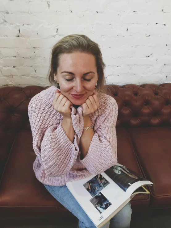 a beautiful blond girl sitting on top of a brown couch holding a magazine