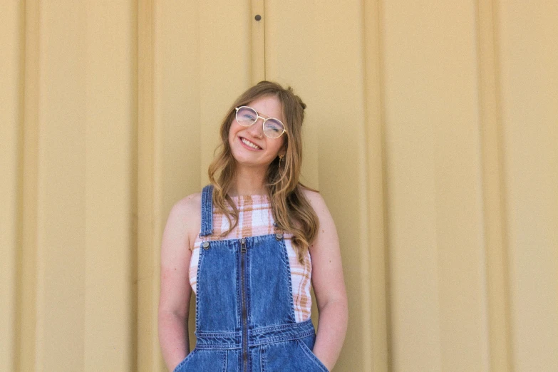 a woman wearing overalls smiles while standing in front of a yellow background