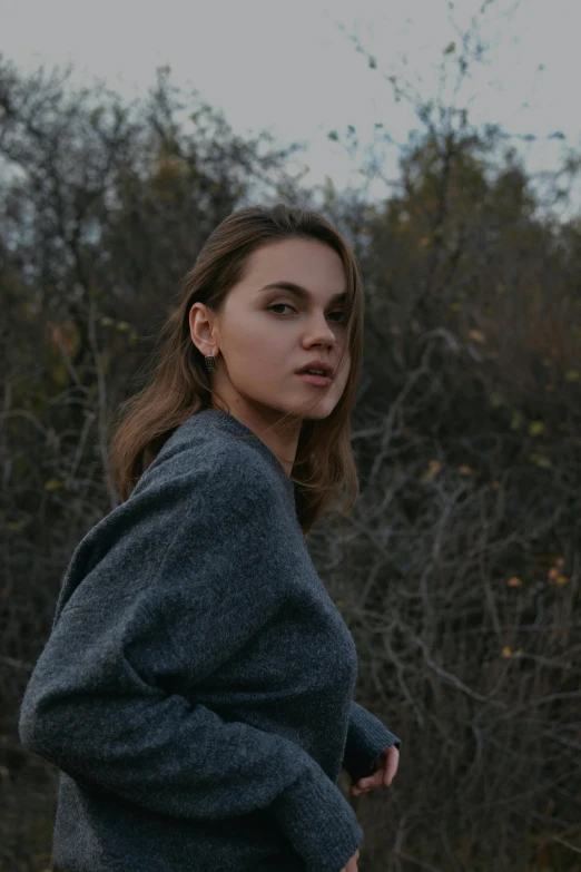 a woman poses in front of tree tops wearing a blue sweater