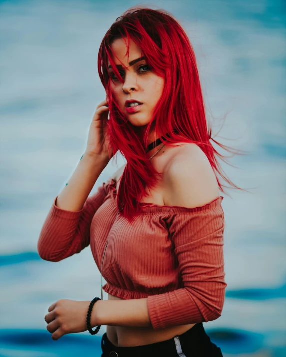 a woman with red hair stands in front of water while wearing a hat