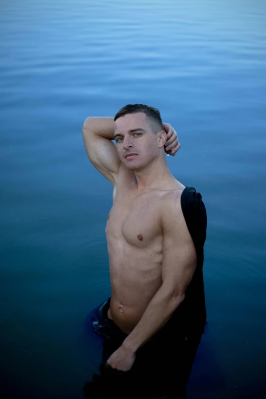 a man poses shirtless in the water with his hand on his shoulder
