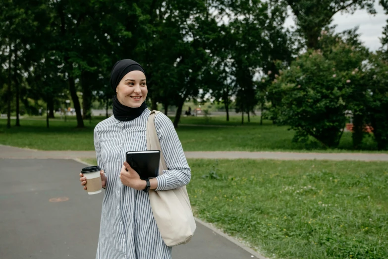 a woman holding a cup and book while walking through a park