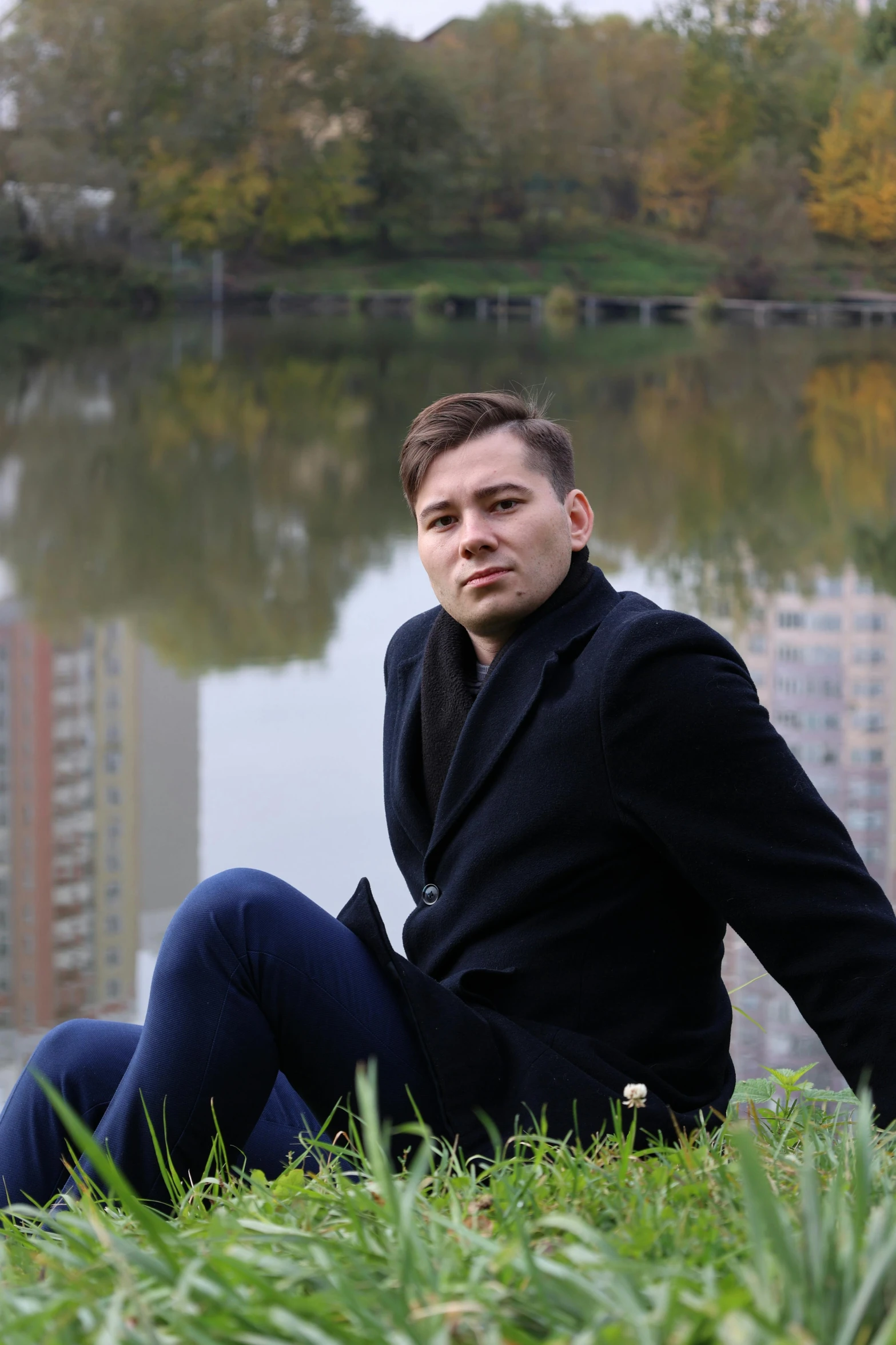 man sitting beside lake in city area with tall buildings