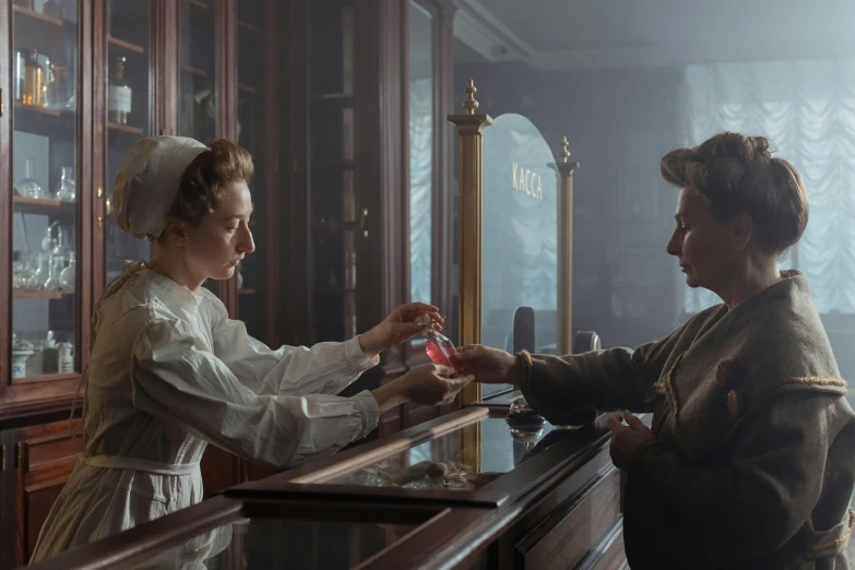 two women in period dresses stand at a counter in front of shelves