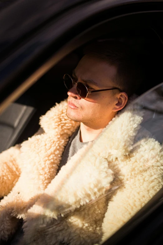 a man wearing sunglasses and a sheary coat looks out the window