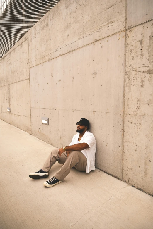 a man sitting against a wall in a white shirt and tan pants