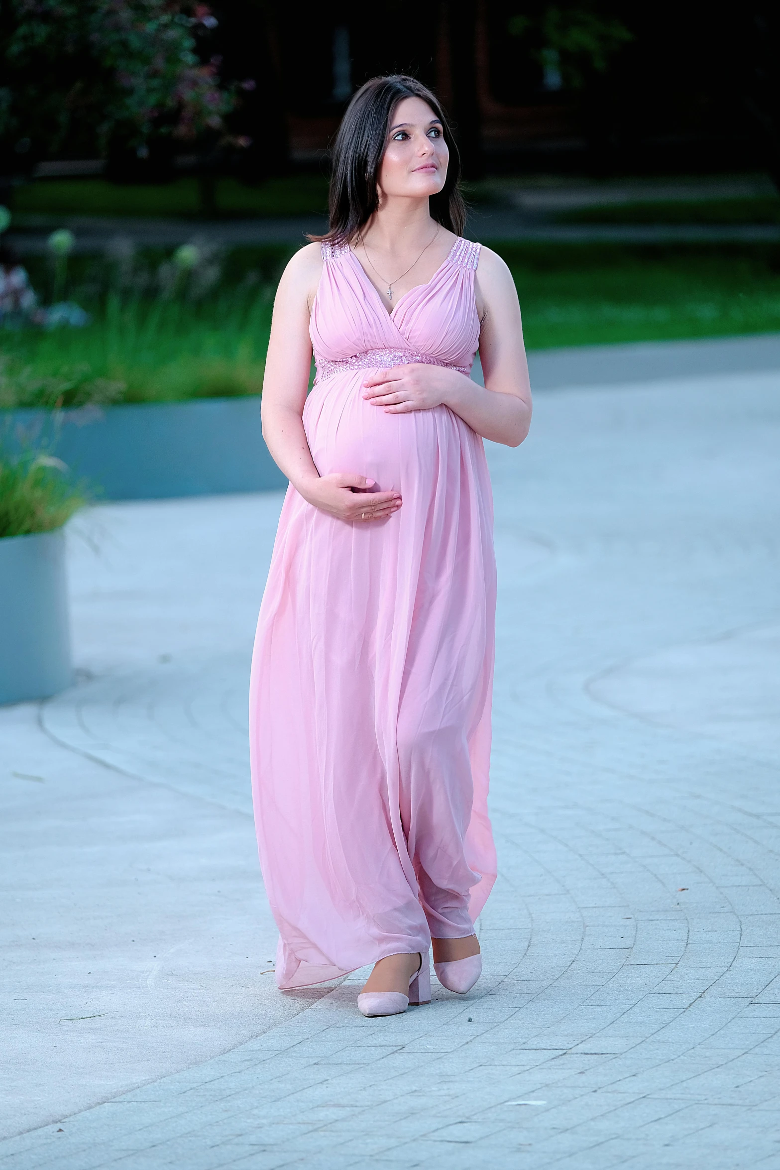 a pregnant woman standing in the middle of a walkway, wearing a pink gown and holding her belly