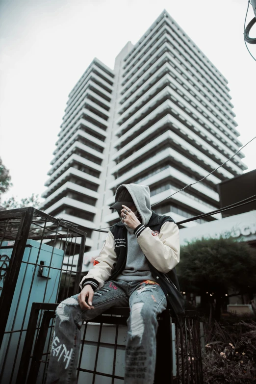 a man wearing a hooded sweatshirt sitting on a fence with a cell phone