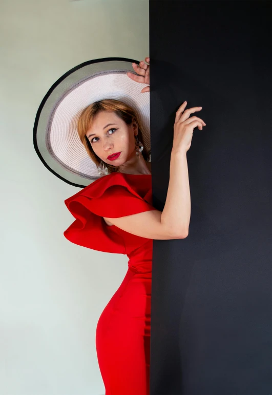 woman in red dress with hat leaning against black wall