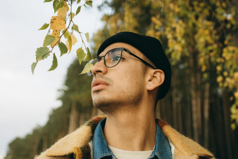 a man in a hat with a coat and glasses is looking up at the leaves on a tree