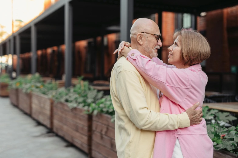 an elderly man and woman hug together in front of an outdoor planter area