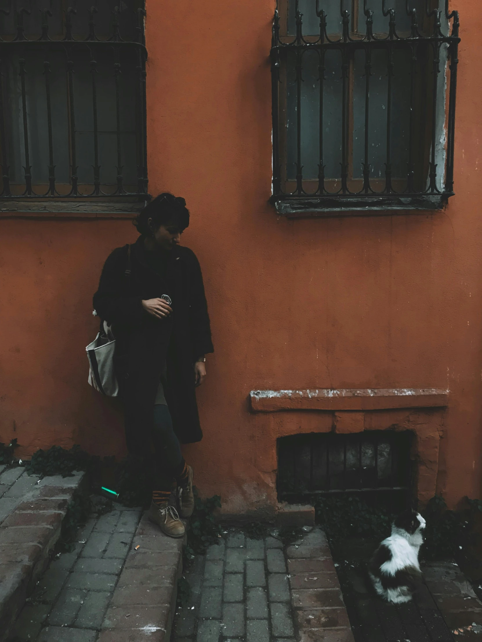 a woman leaning against the wall holding a white purse and looking at her phone