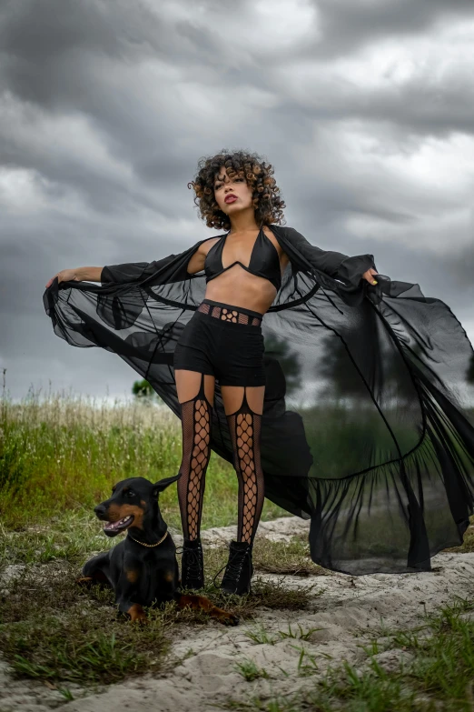 a woman with black stockings, a sheer cape and a dog in a grassy field