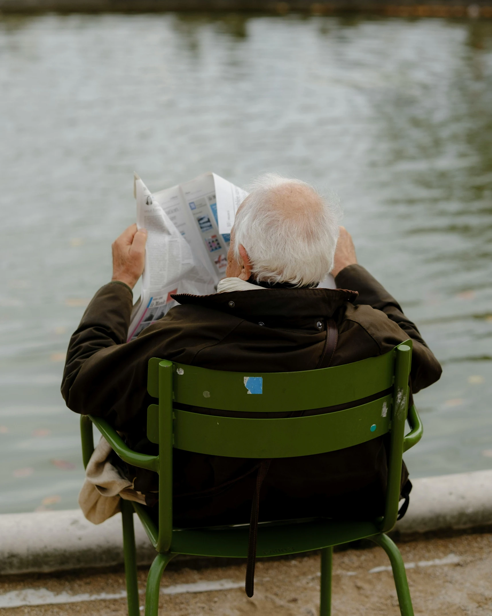 man with white hair, black jacket and greying beard sits on a green lawn chair by the water reading a paper