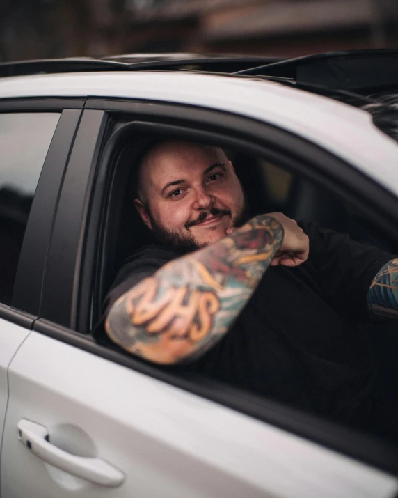a bearded man sitting in his car, with tattoos on his arm