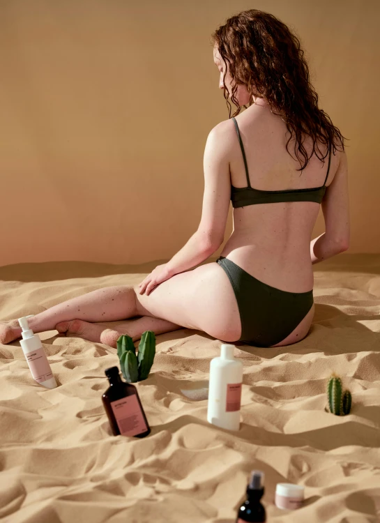 a woman in a bikini sits on the beach and bottles are on the sand