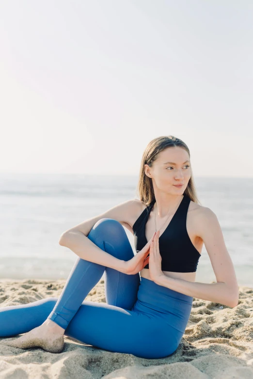 a woman in blue yoga pants and a black top is meditating on a beach