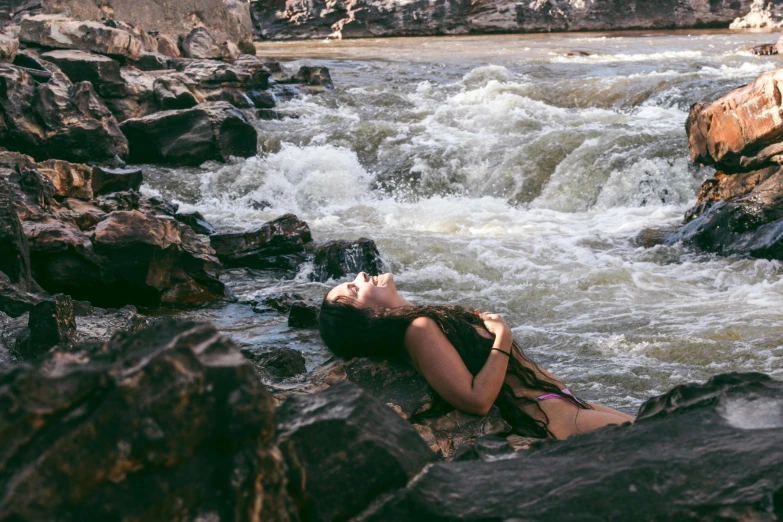 woman laying on rocks near water running past a rocky shore