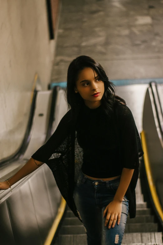 a woman standing on an escalator, wearing blue jeans and black shirt