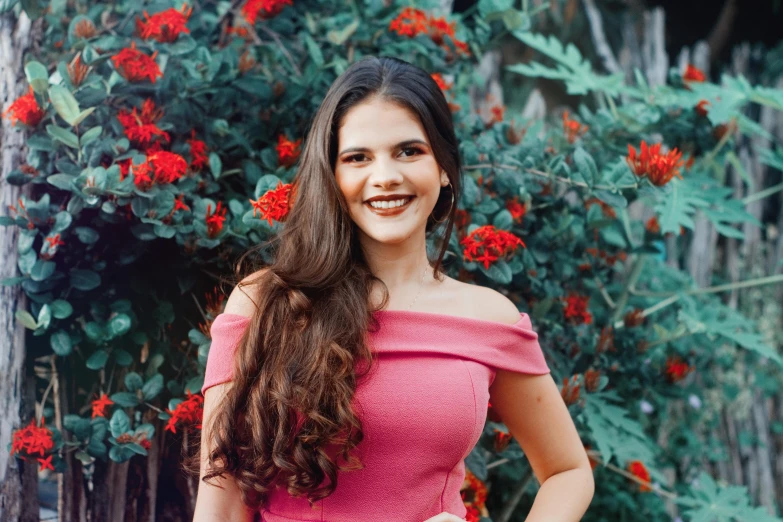 a smiling young woman with long hair wearing pink is standing near a bush