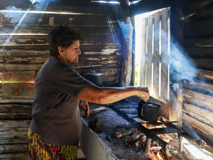 an older woman cooking outside with a log cabin background