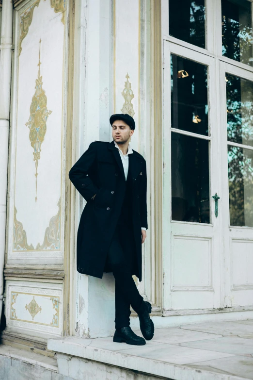 man in coat and fedora leaning on white building