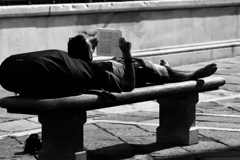 the man is reading a book while lying on a bench