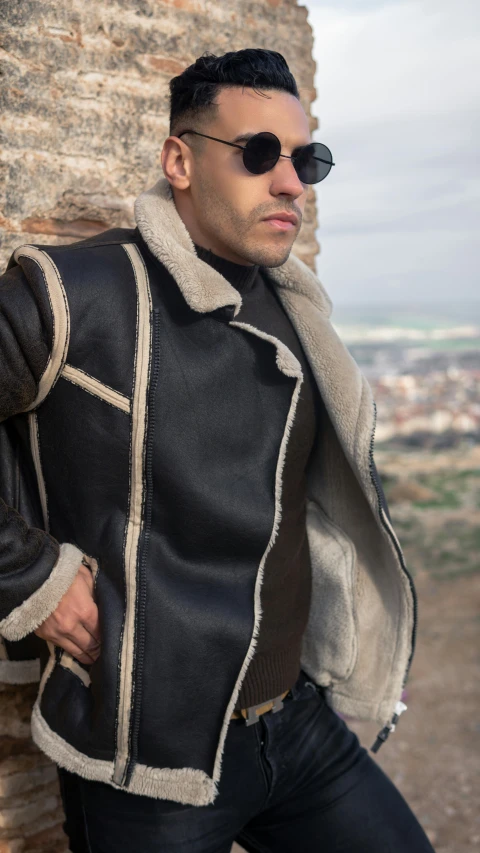 the man in a black jacket is posing by a stone wall