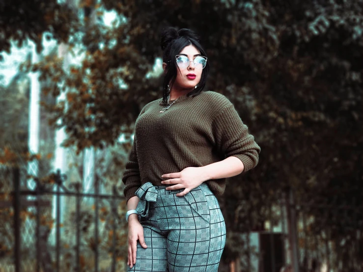 a woman in brown sweater and patterned pants standing by trees