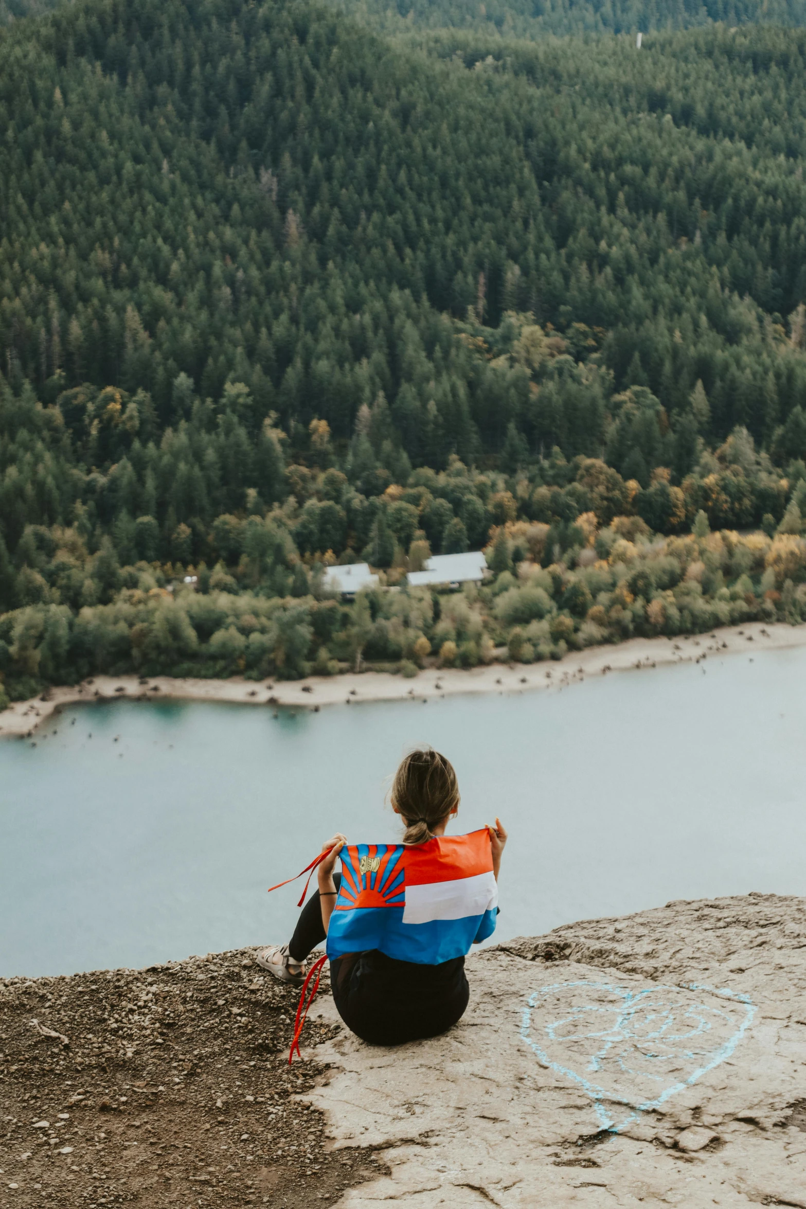 a woman sitting on the edge of a cliff with a tree filled hill in the background