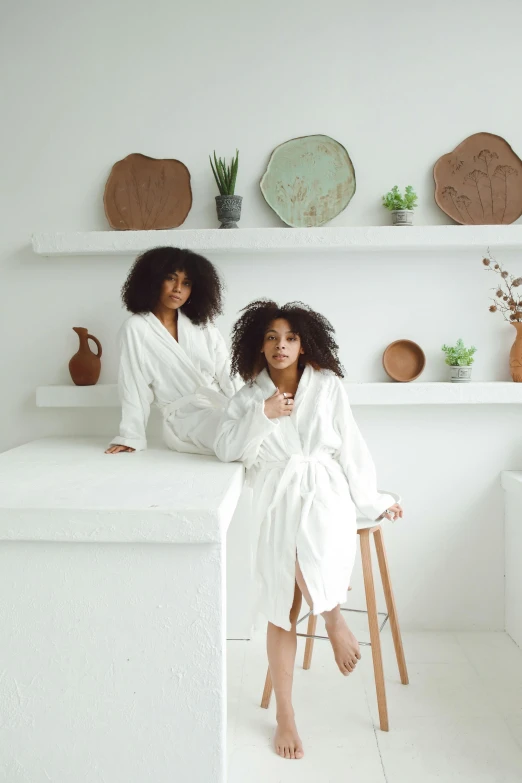 two women in white clothing sitting on stools in a kitchen