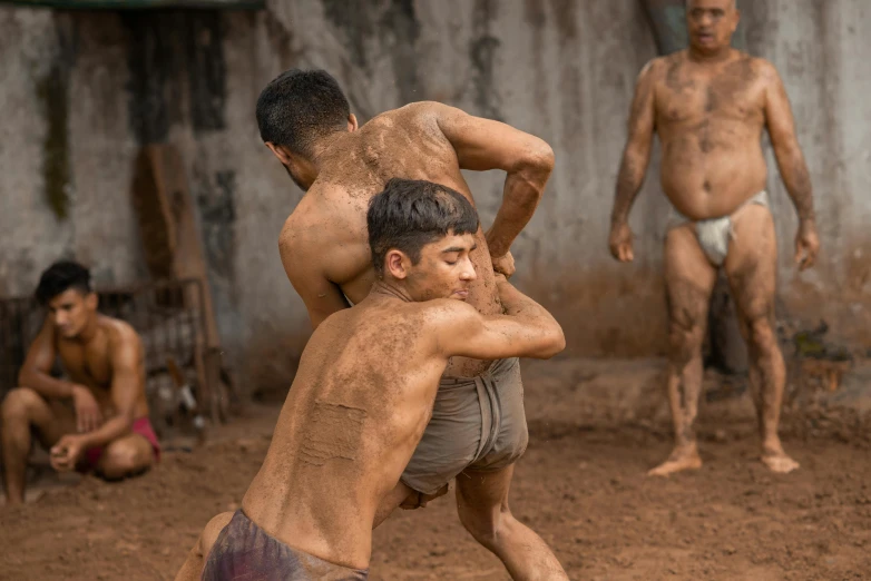 two men, one with no pants and one without shirt, are outside in dirt with mud covering his hands and face