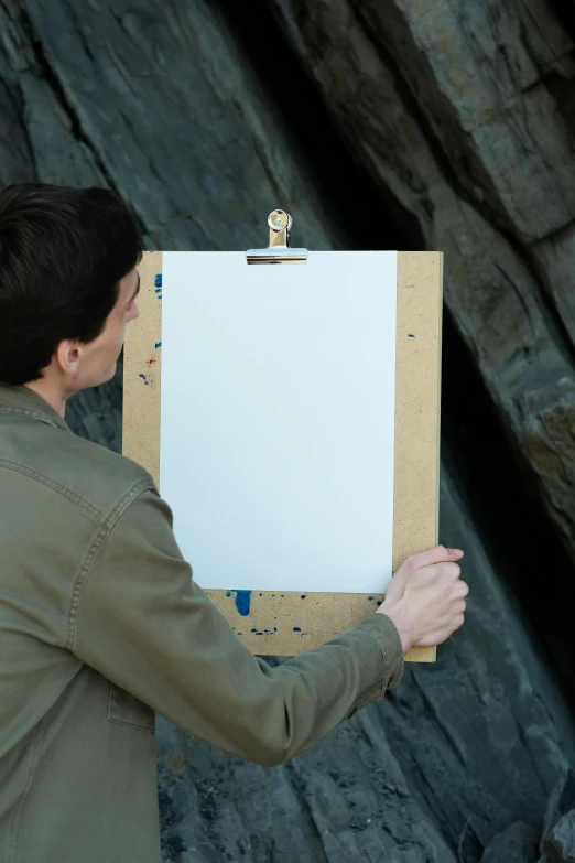 man holding up clipboard on large rock with easel