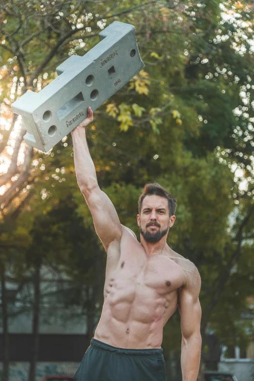 shirtless man holding up a metal box in front of a tree