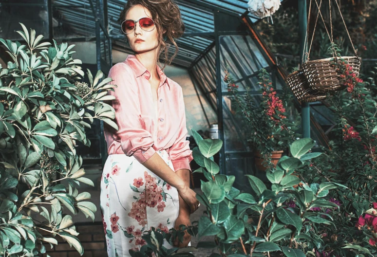 a woman wearing sunglasses leaning against a wall in front of a bunch of plants