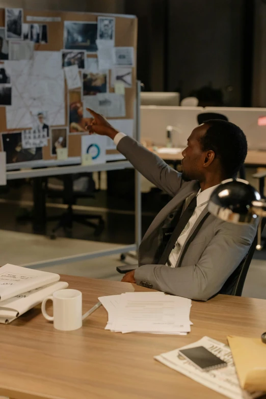 man in suit pointing at bulletin board with papers and laptop on table