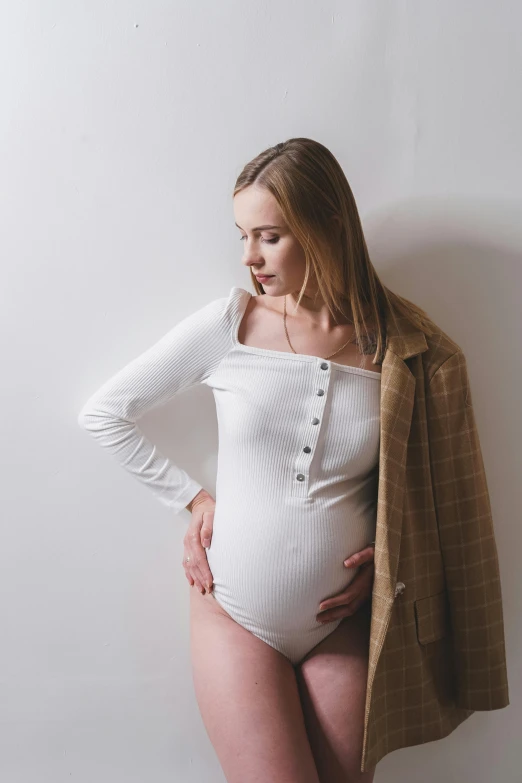 the pregnant woman wears a white bodysuit while standing against a wall