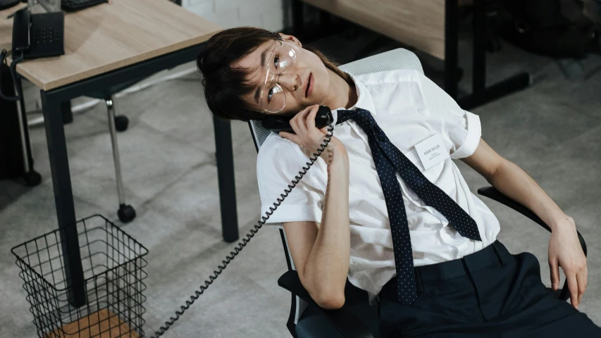 a girl with a tie is sitting at a desk talking on a cell phone